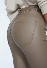Gros plan sur une personne portant un pantalon en cuir synthétique taupe moulant avec des coutures de poches arrière visibles et un haut crème.