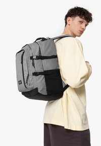 Mochila gris con detalles en negro, múltiples compartimentos y correas ajustables. La tela tiene una apariencia texturizada y duradera.