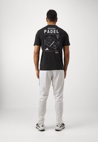 Camiseta de algodón negra con un diseño gráfico en blanco que representa una pista de pádel, texto 'ADIDAS PADEL' y logotipo. Combinada con pantalones blancos y zapatos negros.