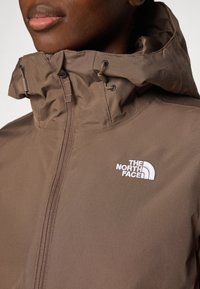 Bruine waterdichte jas met hoge kraag, voorzien van een rits aan de voorkant en witte logo-details. Gladde textuur en verstelbare elementen.