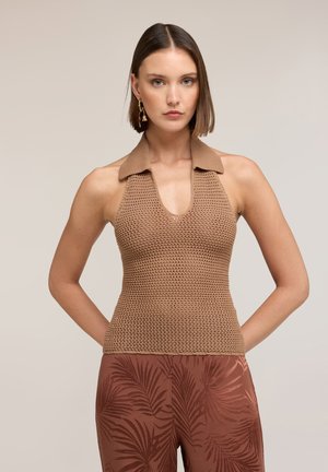 Vrouw met steil bruin haar die een mouwloos gebreid haltertop en een broek met een palmbladpatroon draagt, staand met haar handen achter haar rug.