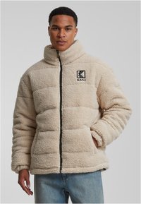 Beige plysj puffer-jakke med høy krage, glidelås foran og store lommer. Har svart logo på brystet og teksturert materiale.