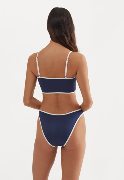 Námořnické modré bikinové sady s bílým lemem. Top typu bustier s nastavitelnými ramínky a vysokými spodními díly, které ukazují hladkou, elastickou texturu.