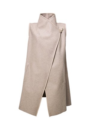 Femme portant un manteau beige sans manches, un pantalon en cuir noir, des talons noirs, tenant un sac à main noir en fourrure, debout devant un fond uni.