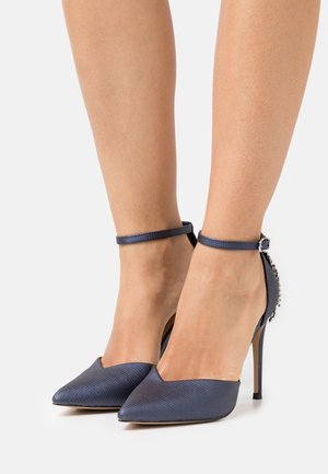 Pieds chaussés de talons hauts pointus bleu marine texturés, avec des brides à la cheville et des embellissements décoratifs à l'arrière.