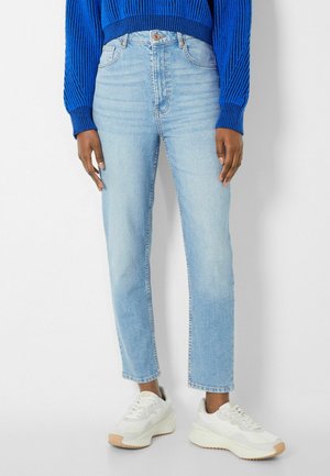 Jeans taille haute bleu clair avec une coupe droite, décoloration subtile et design à cinq poches, associés à des baskets blanches.