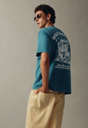Junger Mann mit Sonnenbrille, blauem T-Shirt mit Golfwagen-Design und beigen Hosen, der mit einer Hand in der Tasche steht.