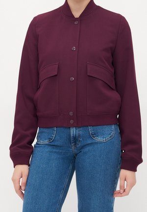 Donna che indossa una giacca color mogano con bottoni e tasche frontali con pattina e jeans blu a vita alta su uno sfondo semplice.
