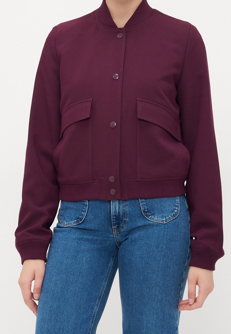 Femme portant une veste bordeaux boutonnée avec des poches à rabat sur le devant et un jean bleu taille haute sur un fond uni.