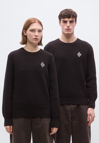 Pulls en maille côtelée brun foncé à manches longues, arborant un petit logo blanc en forme de losange sur la poitrine. Portés avec un pantalon brun assorti.