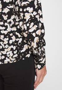 Blouse noire avec un motif abstrait crème et beige, dotée de manches longues froncées et d'un détail bouton au niveau des poignets.