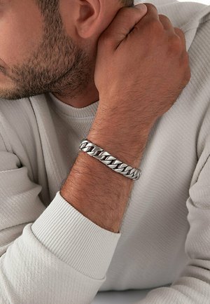 Silver chain bracelet on wrist, featuring thick, interlocking links with a polished surface. Light reflects off the smooth texture.