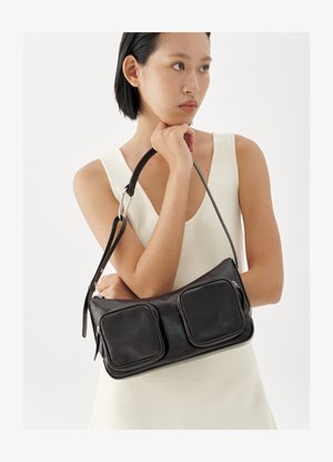 Woman in sleeveless white dress holding black leather handbag with two front zippered pockets against white background.