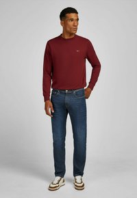 Homme portant un sweat-shirt bordeaux, un jean bleu et des baskets blanches avec des accents marron, se tenant avec une main dans la poche, regardant à droite.