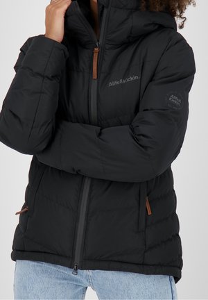 Personne portant une veste matelassée noire avec des poches zippées et une capuche, mains partiellement couvrant le visage, associée à un jean bleu clair.
