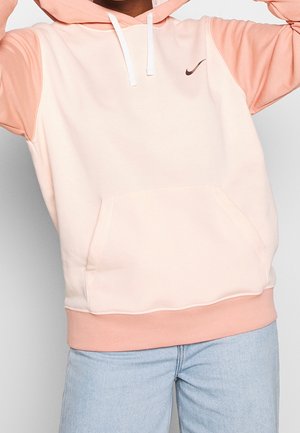 Personne portant un sweat à capuche Nike bicolore pêche et rose clair avec cordons blancs et jean bleu clair, en train de relever la capuche.