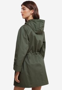 Grøn hættetrøje med en snøret talje, lange ærmer og en glat overflade. Den falder over knæene og har manchetter på ærmerne.