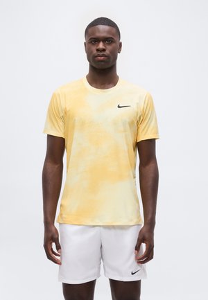 Mand, der står fremad, iført en gul Nike T-shirt og hvide Nike-shorts mod en ensfarvet lys baggrund.