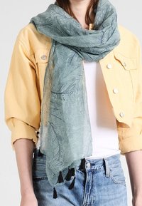 Personne portant une veste courte jaune clair, un haut blanc, un jean bleu et une écharpe bleue à motifs avec des pampilles noires.