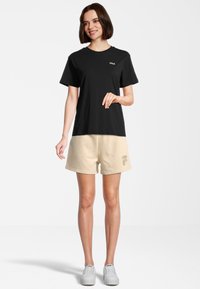 Maglietta nera in cotone con un piccolo logo bianco, abbinata a pantaloni beige con una stampa del logo di lato. Gli shorts hanno una vestibilità morbida e una texture delicata.