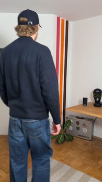 Navy knitted sweater, blue jeans, indoor setting with wooden floor. Wall features orange, pink, and brown stripes; plant and table decor visible.