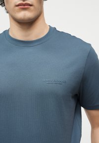Άνδρας φορά μπλε T-shirt Armani Exchange με στρογγυλή λαιμόκοψη και ανάγλυφο λογότυπο "Armani Exchange Milano/New York" στο στήθος.
