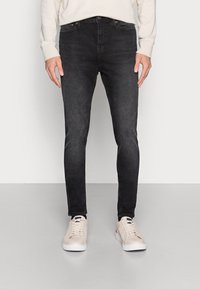 Jeans skinny pretos feitos de denim, com um design cl�ássico de cinco bolsos. Usados com ténis de cor clara sobre um fundo simples.