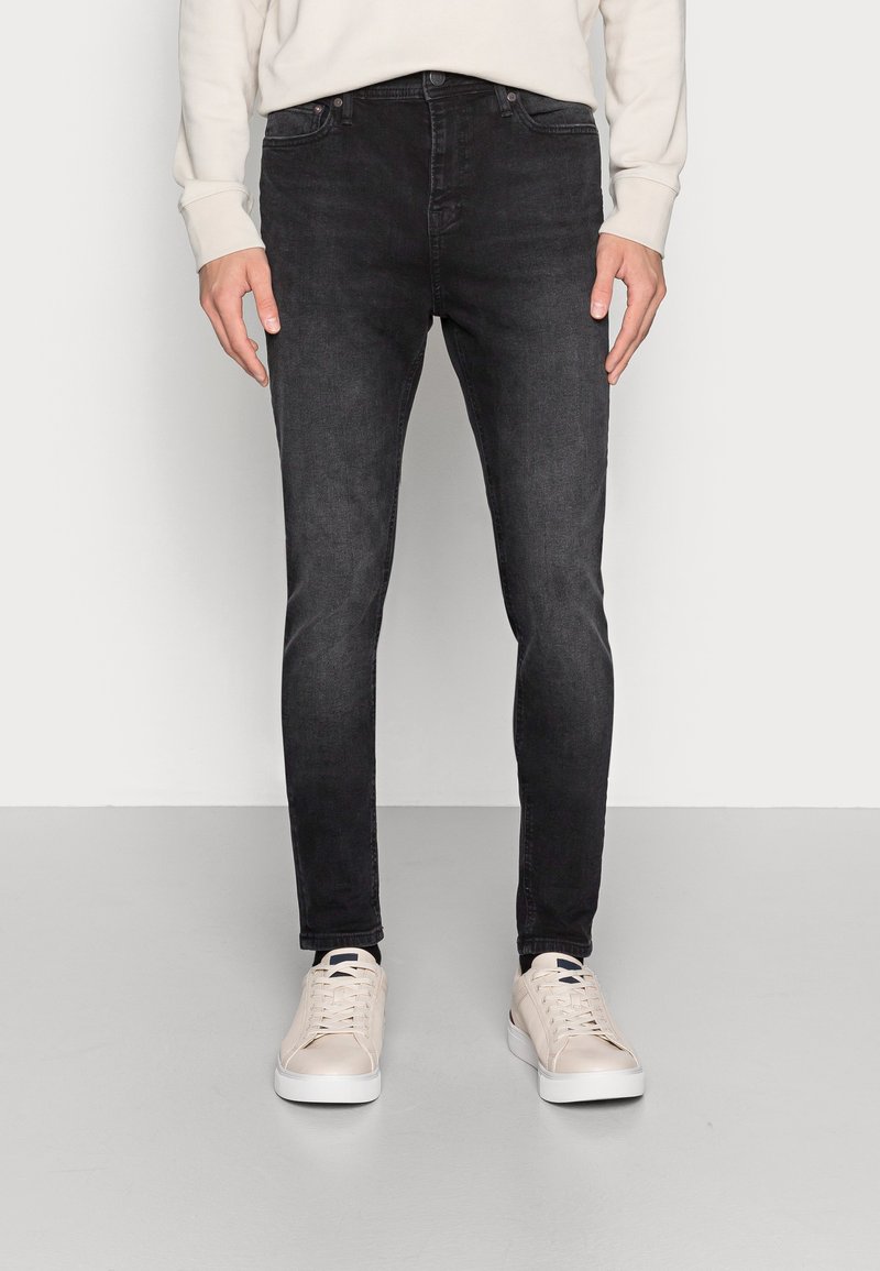 Jeans skinny pretos feitos de denim, com um design clássico de cinco bolsos. Usados com ténis de cor clara sobre um fundo simples.