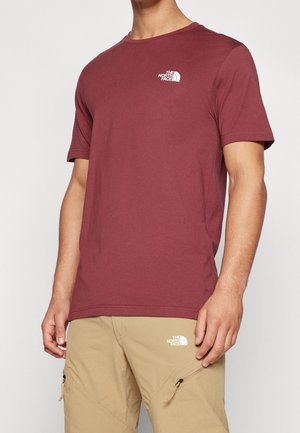 Homme portant un t-shirt bordeaux à manches courtes et un pantalon cargo beige, tous deux avec des logos blancs The North Face sur la poitrine et la cuisse.