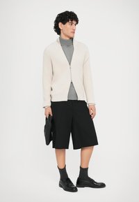 Φωτεινό μπεζ ριμπ πλεκτό ζακέτα με φερμουάρ μπροστά, γκρι ζιβάγκο, μαύρο σορτς και μαύρα παπούτσια, κρατώντας μαύρη τσάντα.