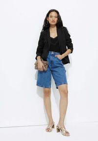 Blazer noir sur un débardeur en dentelle noir, associé à un short en denim bermuda bleu clair. Porte des sandales à talons métalliques et tient une pochette en imprimé léopard.