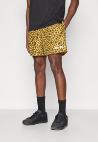 Calções de banho com padrão de leopardo amarelo e textura em malha, apresentando um logótipo branco. Usados com meias pretas e ténis pretos.