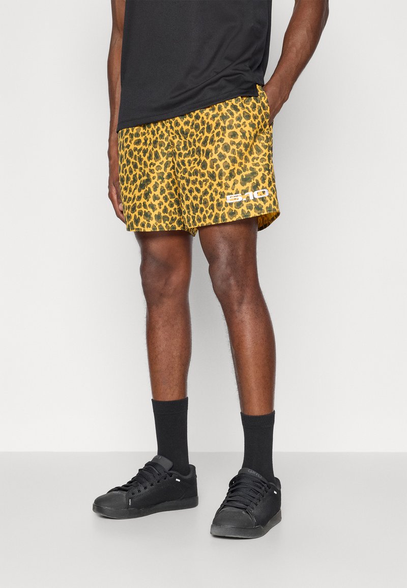 Calções de banho com padrão de leopardo amarelo e textura em malha, apresentando um logótipo branco. Usados com meias pretas e ténis pretos.