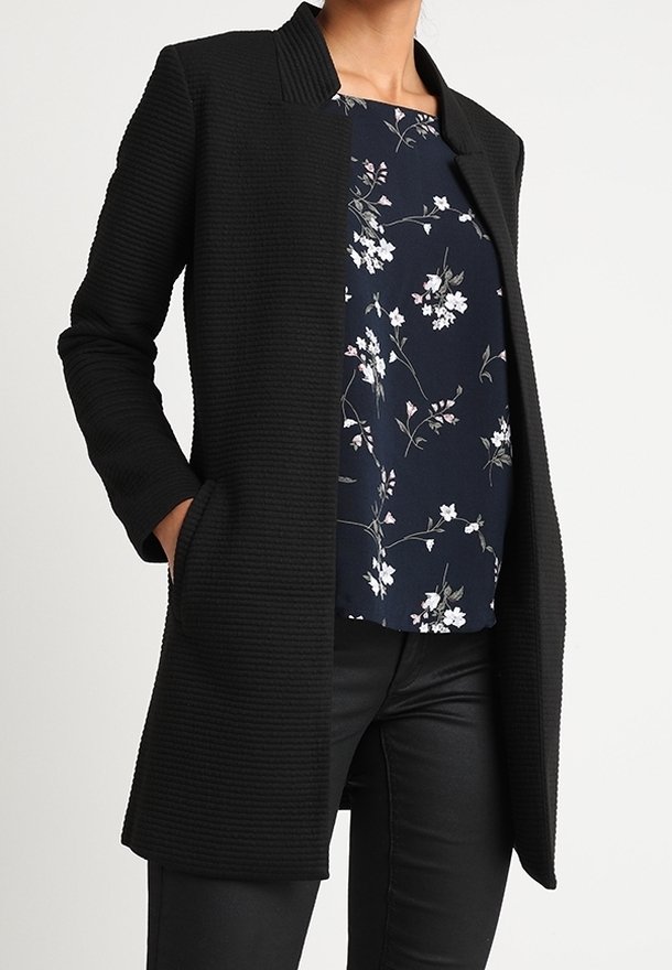 Manteau noir texturé avec un col, des poches avant et une coupe ouverte. Porté sur un haut à motifs floraux et associé à un pantalon foncé.
