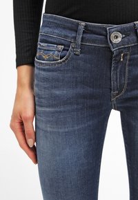 Jeans de denim azul oscuro con un ajuste slim, que cuentan con bolsillos frontales con costura decorativa y un cierre de botón superior plateado. Textura suave.