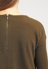 Suéter verde oliva con una textura suave, que cuenta con un detalle de cremallera en la parte posterior del escote para una fácil colocación.