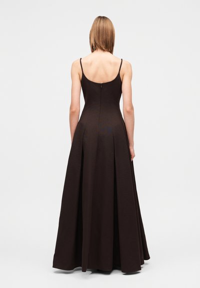 Mulher de costas, vestindo um vestido castanho escuro sem mangas, comprido, com alças finas e fecho de correr nas costas.