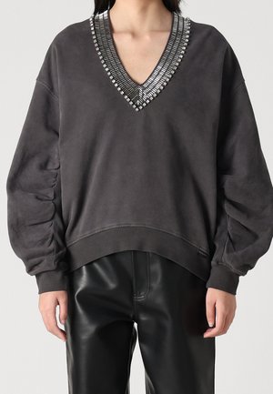 Žena nosící tmavě šedý svetr s dlouhým rukávem s nařasenými rukávy a výstřihem do V s korálky, ve spojení s černými koženými kalhotami.