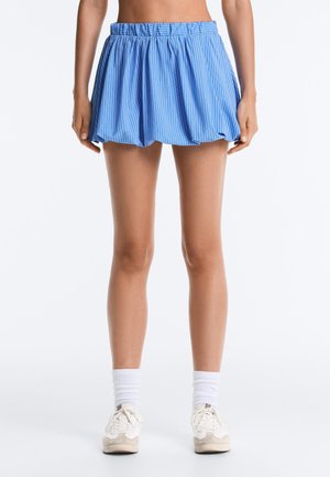 Žena nosiaca voľnú modrobielo pruhovanú mini sukňu s elastickým pásom, biele členkové ponožky a svetlé tenisky, stojaca vzpriamene.