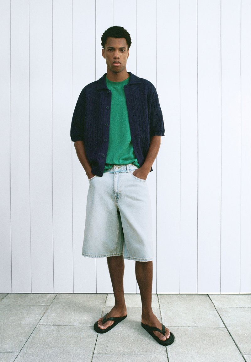 Jeune homme debout les mains dans les poches, portant une chemise verte, un cardigan bleu foncé à manches courtes, un short en jean clair et des tongs noires.