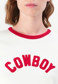 Valkoinen t-paita, jossa on punaiset ohutraita-aukot ja sana "COWBOY" paksuina, brodeerattuina punaisina kirjaimina keskitettynä rinnassa. Sujuva puuvillapinta.