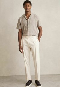 Helles graues gemustertes Kurzarmhemd, cremefarbene maßgeschneiderte Hose und schwarze Sneaker. Glattes Material und klare Linien. Knopfleiste.