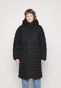 Zwarte pufferjas met een gestructureerd, golvend oppervlak, kap en trekkoord. Voorzien van een volledige rits en zijzakken. Gecombineerd met blauwe jeans.