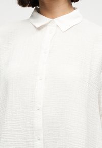 Λευκό μακρυμάνικο πουκάμισο με υφή, κουμπιά κατά μήκος του μπροστινού μέρους και κλασικό μυτερό γιακά, που φοριέται από άτομο όπου φαίνεται μόνο ο λαιμός.