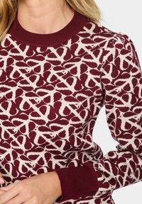 Pull en tricot bordeaux présentant un motif abstrait crème, un col rond côtelé, des poignets avec manches légèrement bouffantes. Texture douce.
