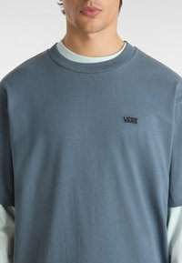 Camiseta de algodón azul claro con cuello redondo y mangas cortas. Presenta un pequeño logo negro de Vans en el lado izquierdo del pecho. Textura suave.