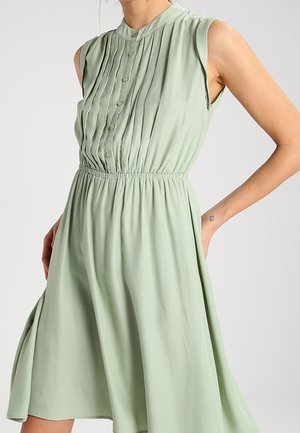 Mouwloze lichtgroene jurk met geplooide kraag met knopen en elastische tailleband, met uitlopende rok, gedragen door persoon met hand in de zak.