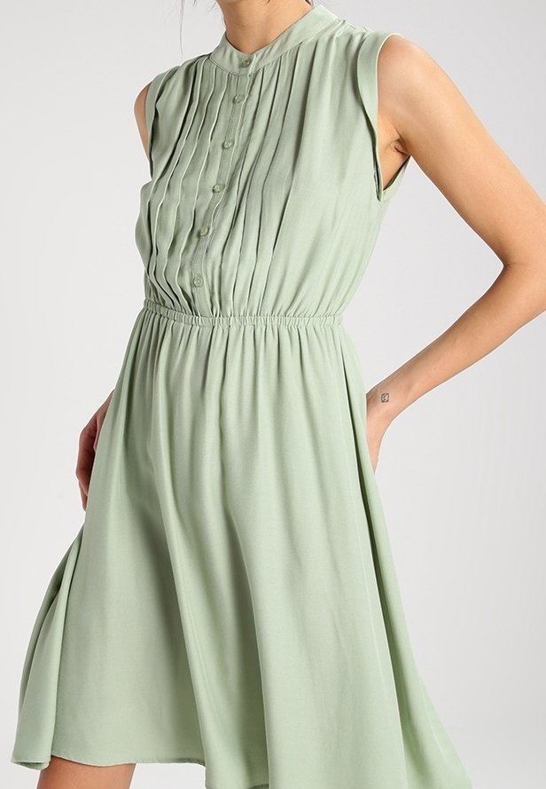 Robe sans manches vert pâle avec corsage plissé boutonné et ceinture élastique, jupe fluide, portée par une personne avec la main dans la poche.