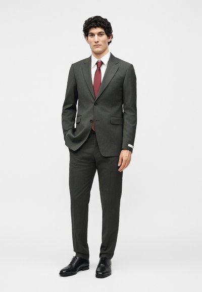 Άνδρας με σγουρά μαλλιά που φοράει σκούρο πράσινο κοστούμι, λευκό πουκάμισο, μπορντό γραβάτα και μαύρα επίσημα παπούτσια, στέκεται με το ένα χέρι στην τσέπη.