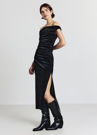 Schwarzes schulterfreies Kleid mit geraffter Textur, seitlichem Schlitz und figurbetonter Silhouette. Kombiniert mit hohen schwarzen Lederstiefeln und minimalem Schmuck.